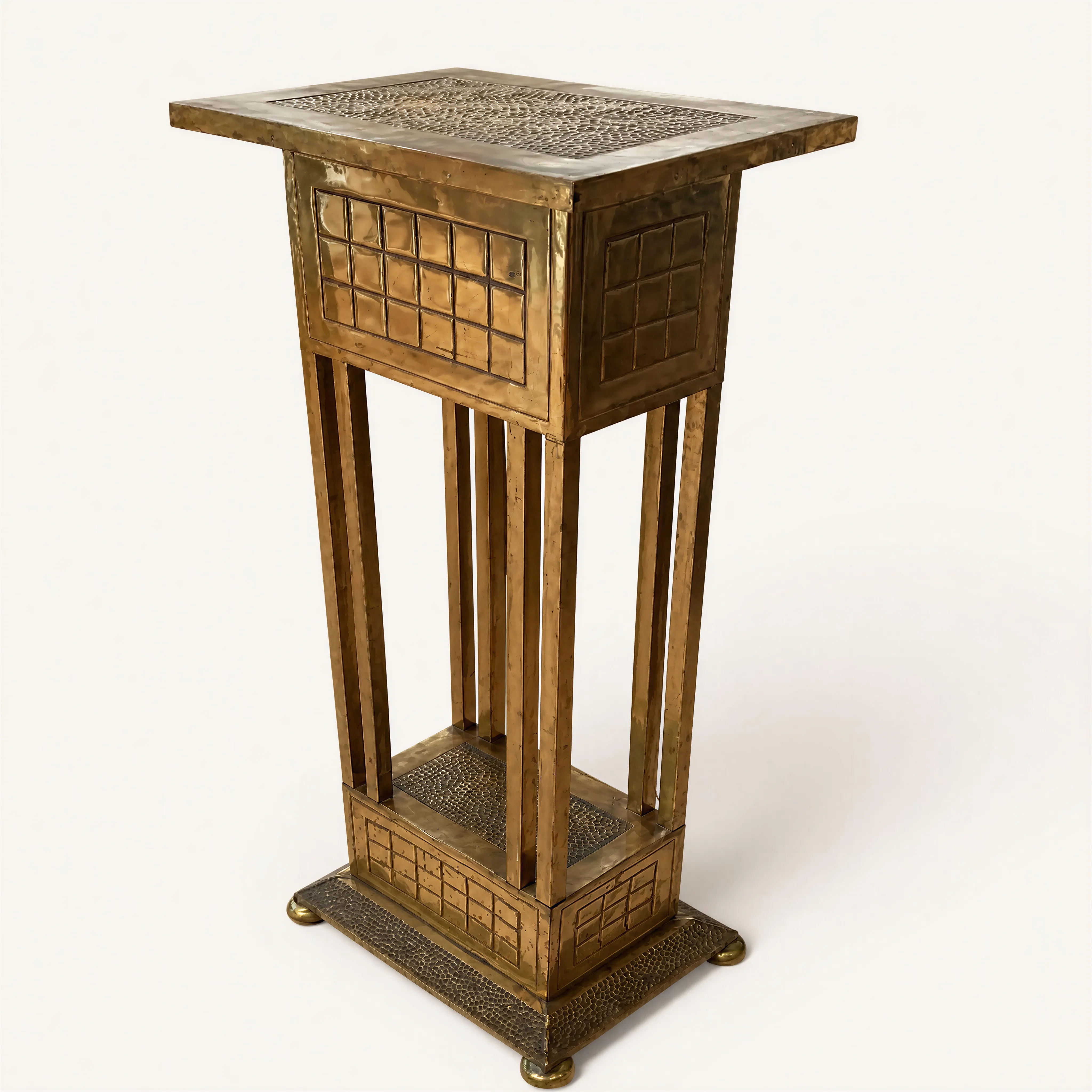 Sellette de présentation de forme architecturale et géométrique. La structure en bois est intégralement habillée de plaques de laiton. Elle se compose d'un plateau supérieur rectangulaire débordant et d'une base pleine, tous deux agrémentés en leur centre d'une surface texturée au marteau. Les blocs supérieur et inférieur présentent un décor quadrillé constitué de carrés repoussés en léger relief. Ces deux registres sont reliés par quatre montants verticaux et droits. L'ensemble repose sur quatre petits pieds de forme sphérique.