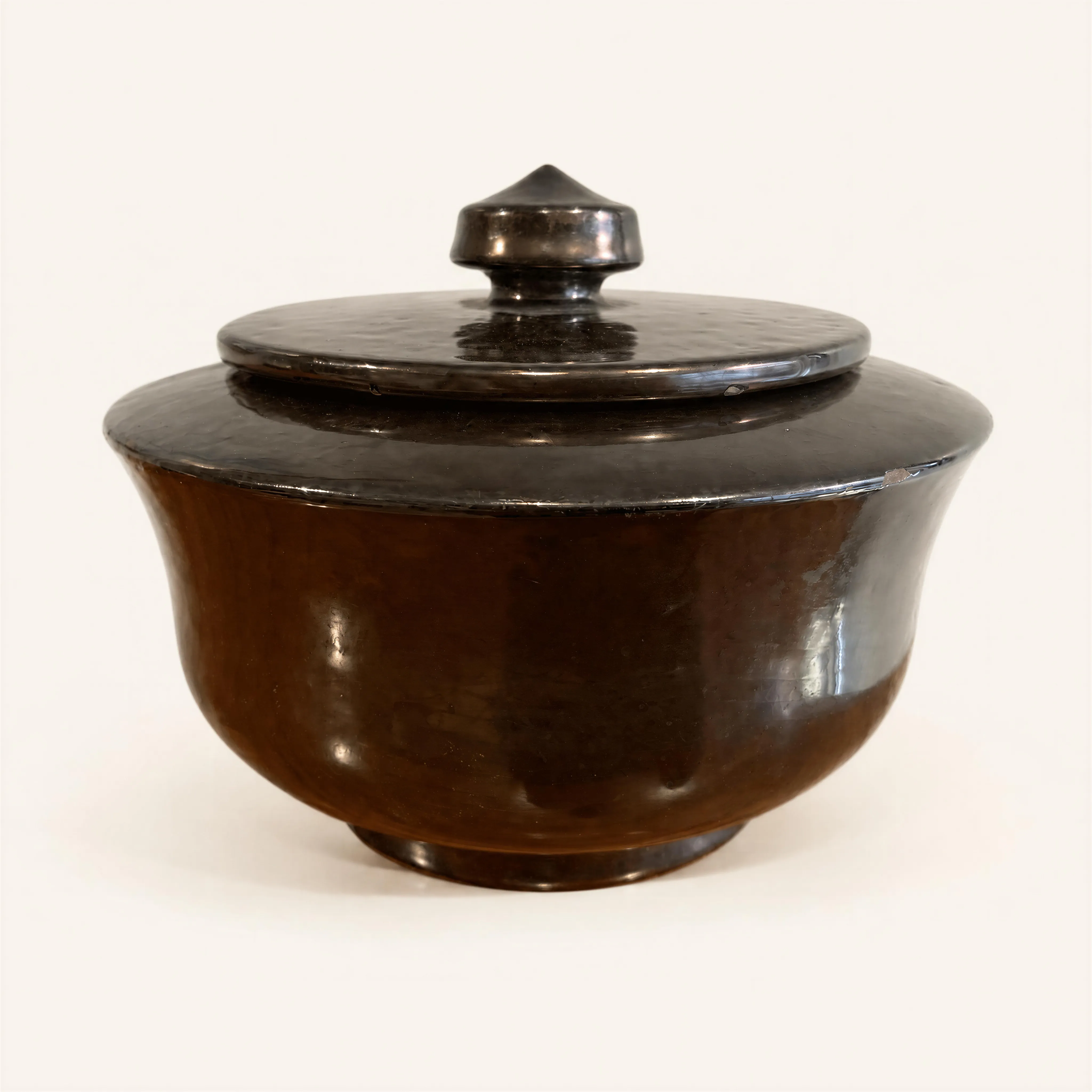 Covered circular ceramic pot. The bulbous body flares upward before narrowing at the rim, resting on a recessed ring base. The piece features a flat, fitted lid topped in its center by a tiered, conical knob handle. The exterior is finished with a glossy black luster glaze.