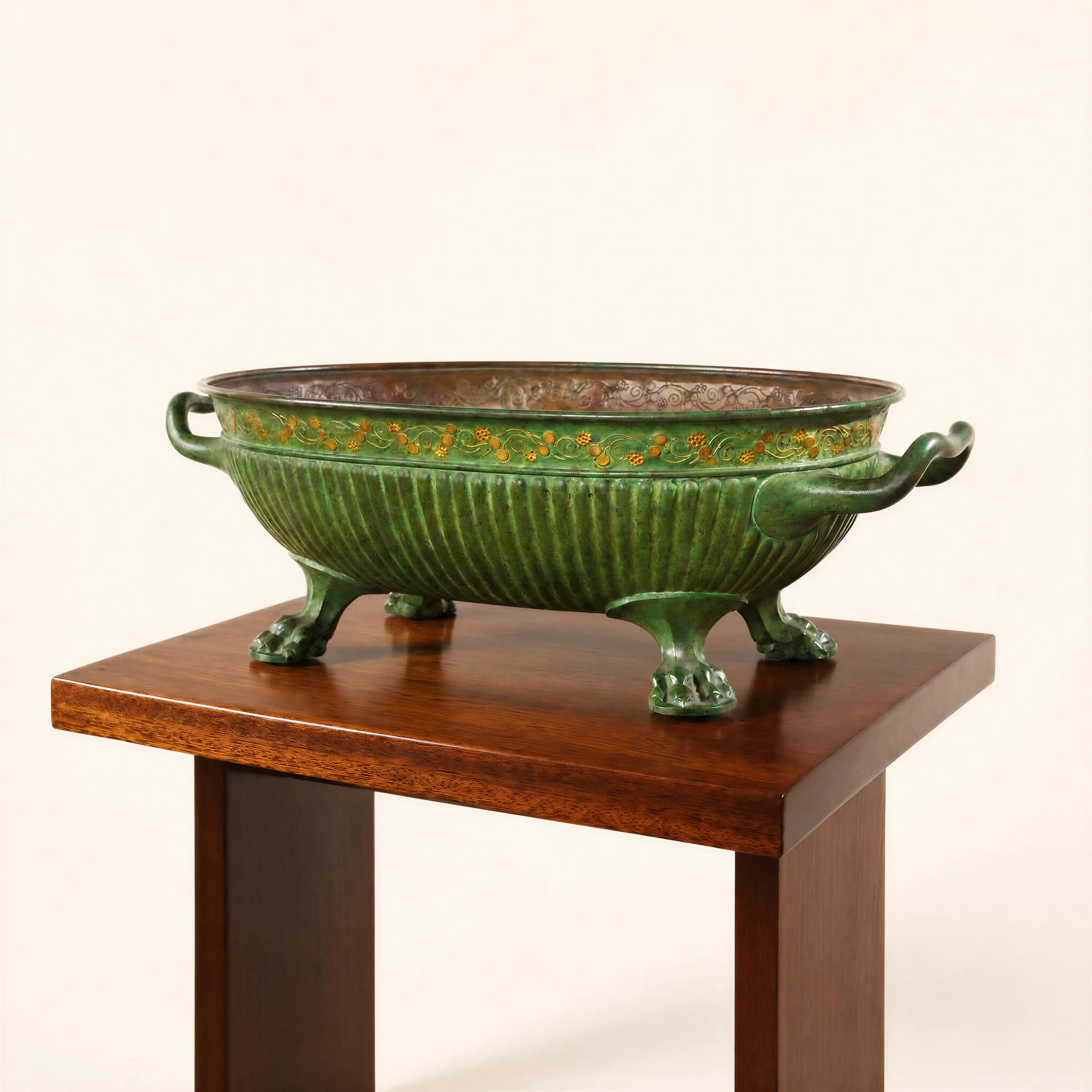 Large oval-shaped bronze and brass bowl, resting on four sturdy antique-inspired lion paw feet. The lower body is richly crafted with a fluted relief (vertical ribs), highlighting the elegance of its volume. The upper section, just below the rim, is adorned with a delicate inlaid frieze of golden brass scrolls and floral motifs, which stand out against the deep, textured green patina covering the exterior of the artwork. Two side handles with sinuous curves complete this sculptural composition.
Signed and crafted between 1909 and 1913.