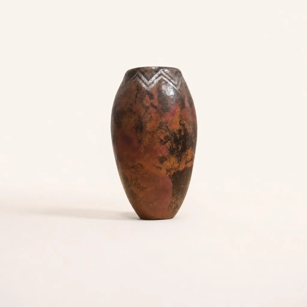 An elongated ovoid dinanderie vase. The narrowed neck is highlighted by a delicate geometric frieze of silvered zigzags (or chevrons), above a body adorned with a rich mottled red, brown, and copper fire-patina. 

Signed "CL Linossier 1927”.