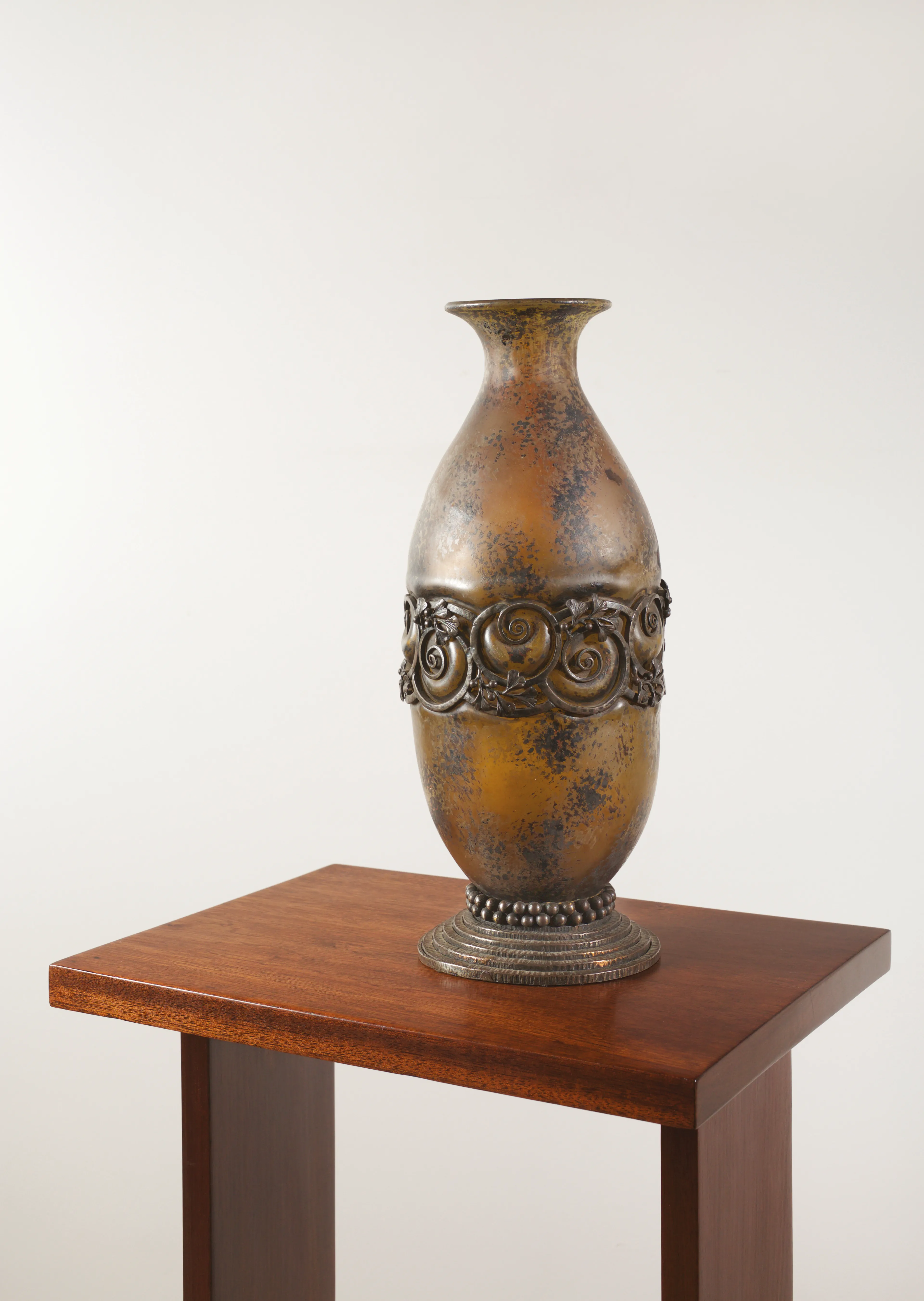 An elongated ovoid vase with a flared neck made of Daum Frères glass blown into its wrought-iron mount of Brandt. The mottled intercalary glass body features rich amber, golden, and deep brown inclusions. The middle of the body is encircled by a wide, openwork metallic band decorated with a continuous, elegant frieze of spiraling volutes interwoven with stylized foliage and berries. The vessel rests on a circular pedestal foot highlighted by a row of wrought-iron beads, sitting atop a stepped base with concentric rings.
Signed "Daum" and "Brandt".
