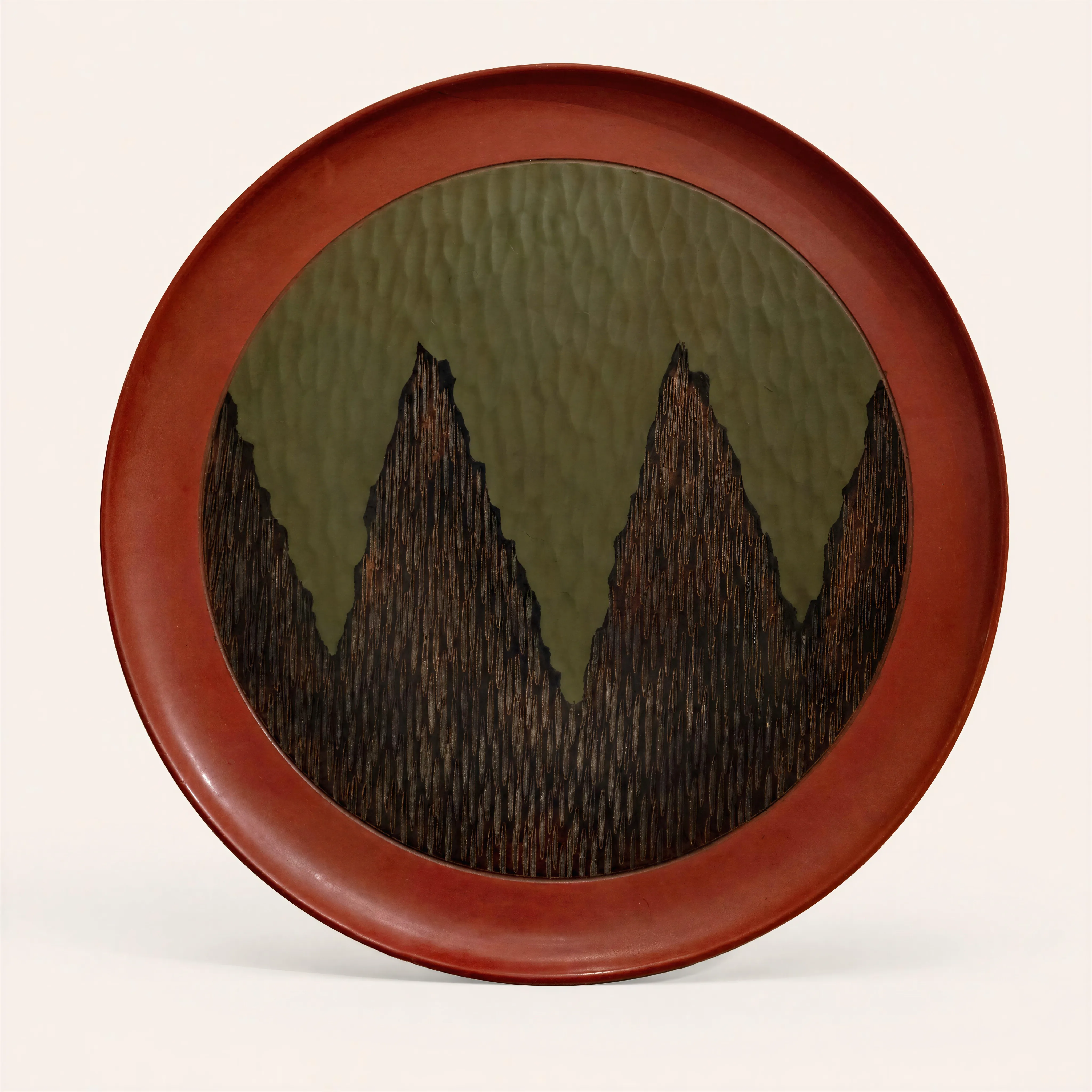 Circular lacquered wood plate. The central part is decorated with a rich textured design featuring three large pointed motifs, evoking dark, striated mountain peaks. These jagged silhouettes stand out against an olive green background, which is also finely striated. The wide rim of the plate features a smooth brick-red lacquer, offering a sharp contrast to the dark colors and textured relief of the center.  