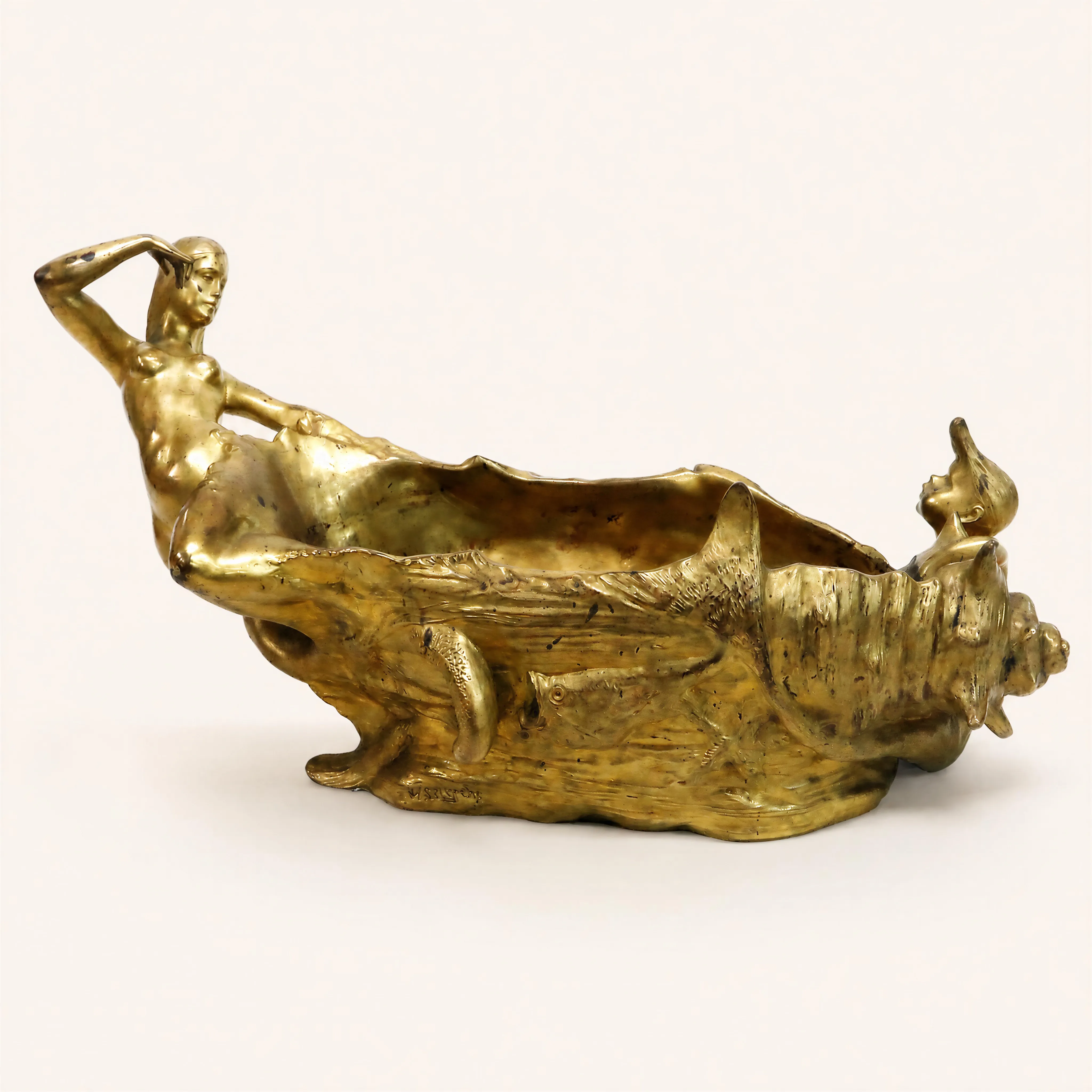 Jardinière de style Art Nouveau en bronze doré, sculptée d’un décor marin en relief. La structure organique, évoquant le mouvement des vagues, met en scène deux figures sculptées aux extrémités : d'un côté, une nymphe dénudée allongée sur la lèvre du vase, le bras droit replié sur son visage ; de l'autre, un jeune enfant émergeant d'un grand coquillage en forme de conque. Les flancs de la pièce sont agrémentés de motifs ciselés de poissons et d'une queue marine en relief.
Signé « HSt Lerche » sur la base
