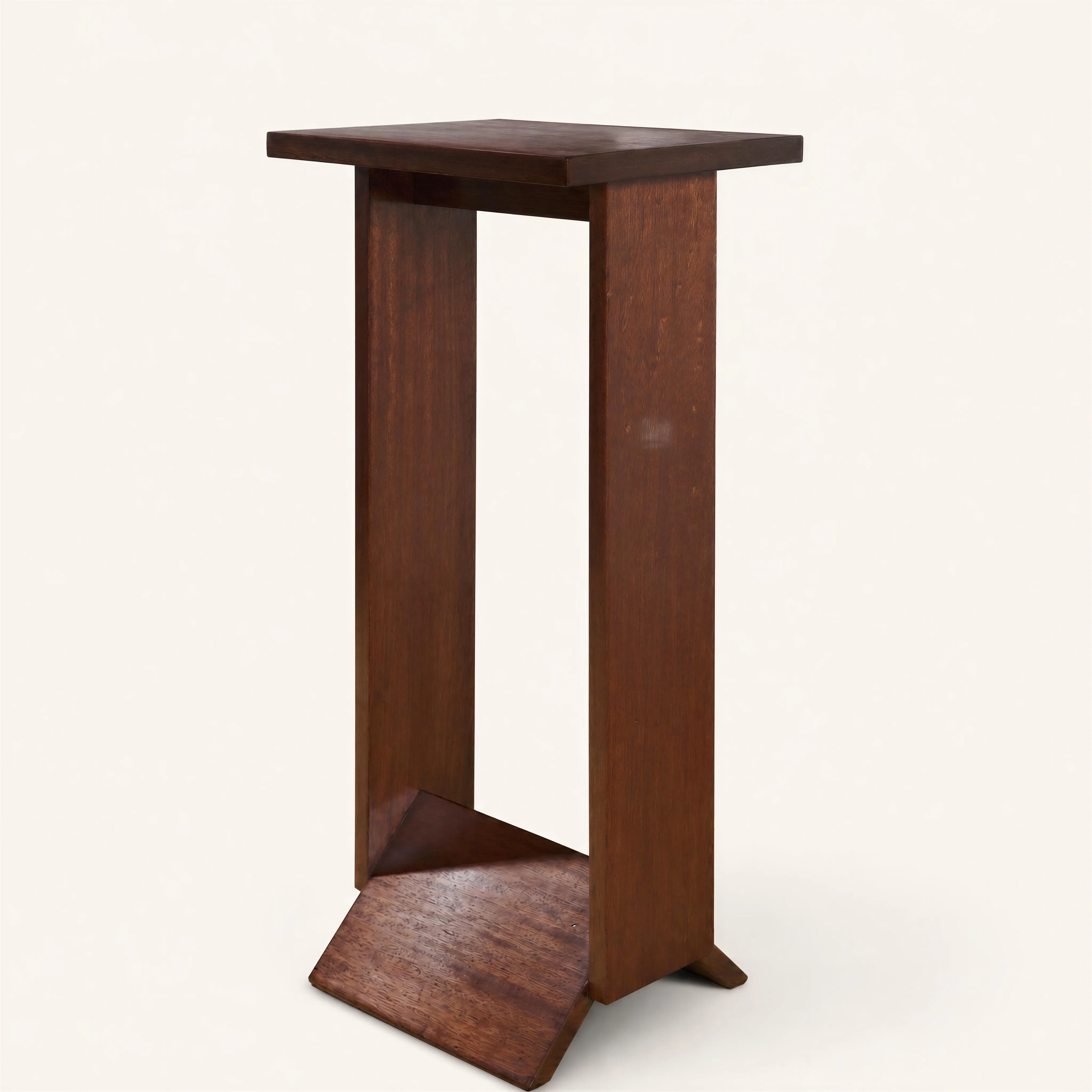 Table haute d'appoint au design profondément moderniste et architectural, caractéristique du travail de Pierre Chareau. La pièce est construite à partir de larges pans de bois francs et géométriques en acajou, mettant en valeur la chaleur et le veinage naturel du matériau.  
Le plateau supérieur, plat et rectangulaire, repose sur deux solides montants verticaux pleins. La base est stabilisée par deux plans inclinés formant un « V » inversé (ou un livre ouvert), complétés par de discrets patins débordant à la base des montants. L'ensemble s'impose par ses lignes rigoureuses, totalement dépourvues d'ornementation superflue.