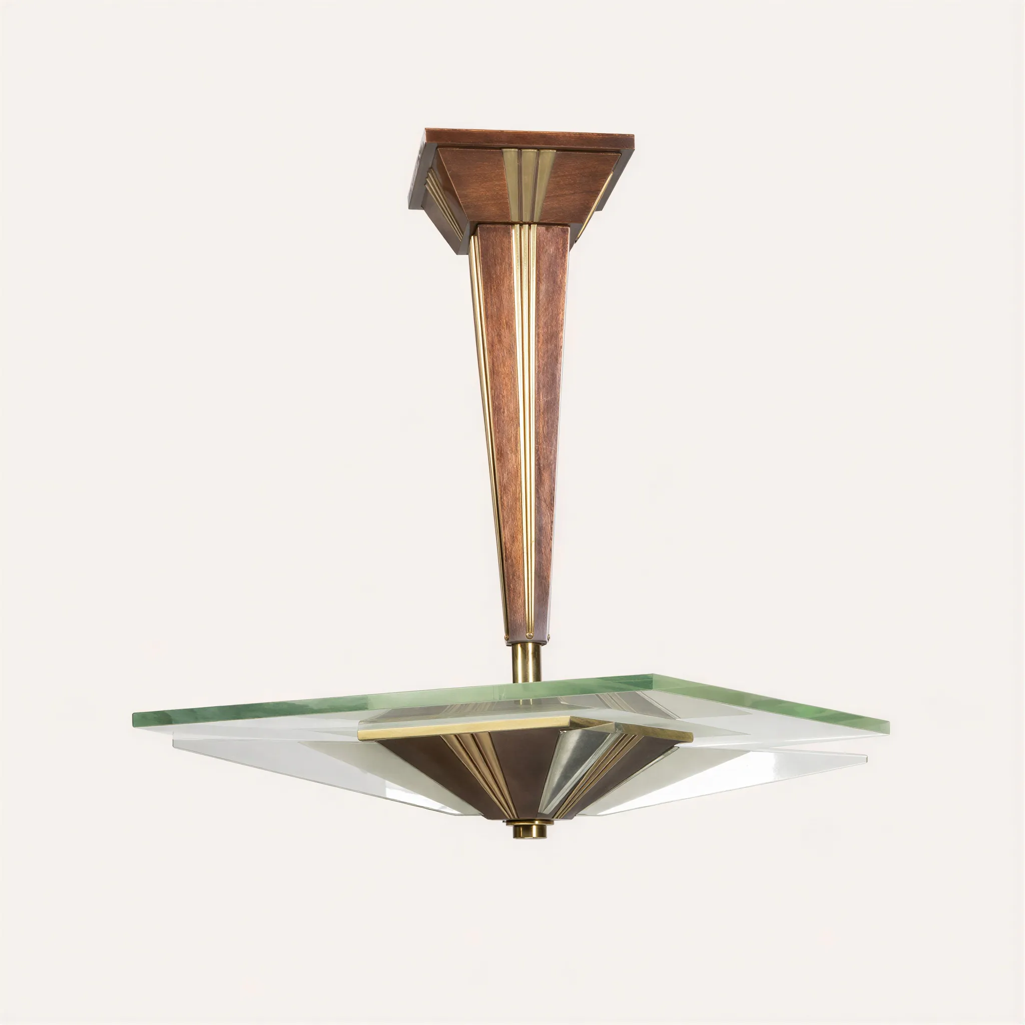 Lustre moderniste à monture géométrique en acajou et métal. La monture centrale prend la forme d'un long fût en acajou de section pyramidale inversée, rehaussé de bandes de métal aux motifs striés verticaux. Cette tige soutient une large dalle carrée en verre translucide, dont les quatre angles sont agrémentés de biseaux en verre. Le luminaire se termine dans sa partie inférieure par un cache-bélière en pointe inversée assorti, mêlant acajou et métal.