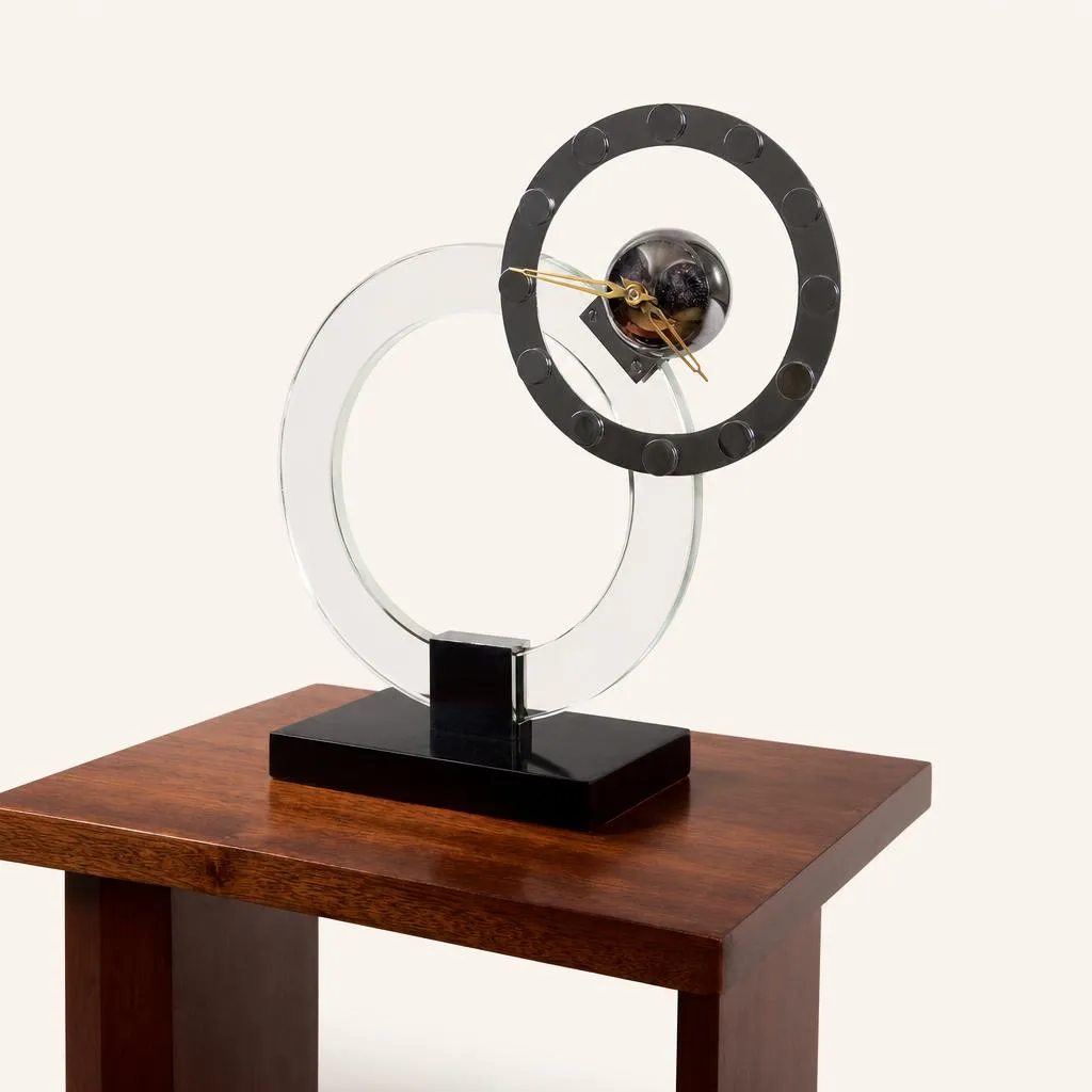 This modernist clock features a geometric composition structured around two overlapping rings. A large, thick translucent glass ring rises vertically from a rectangular black lacquered wooden base. Intersecting it, a smaller chromed metal ring serving as the dial is adorned with raised circular hour markers and encloses a central metal sphere housing the movement and hands.

Literature:
• “Art et Décoration”, 1932, page 322.  
• Alain-René Hardy, “Jacques Adnet”, Les Éditions de l'Amateur, Paris, 2009, reproduced on page 203.