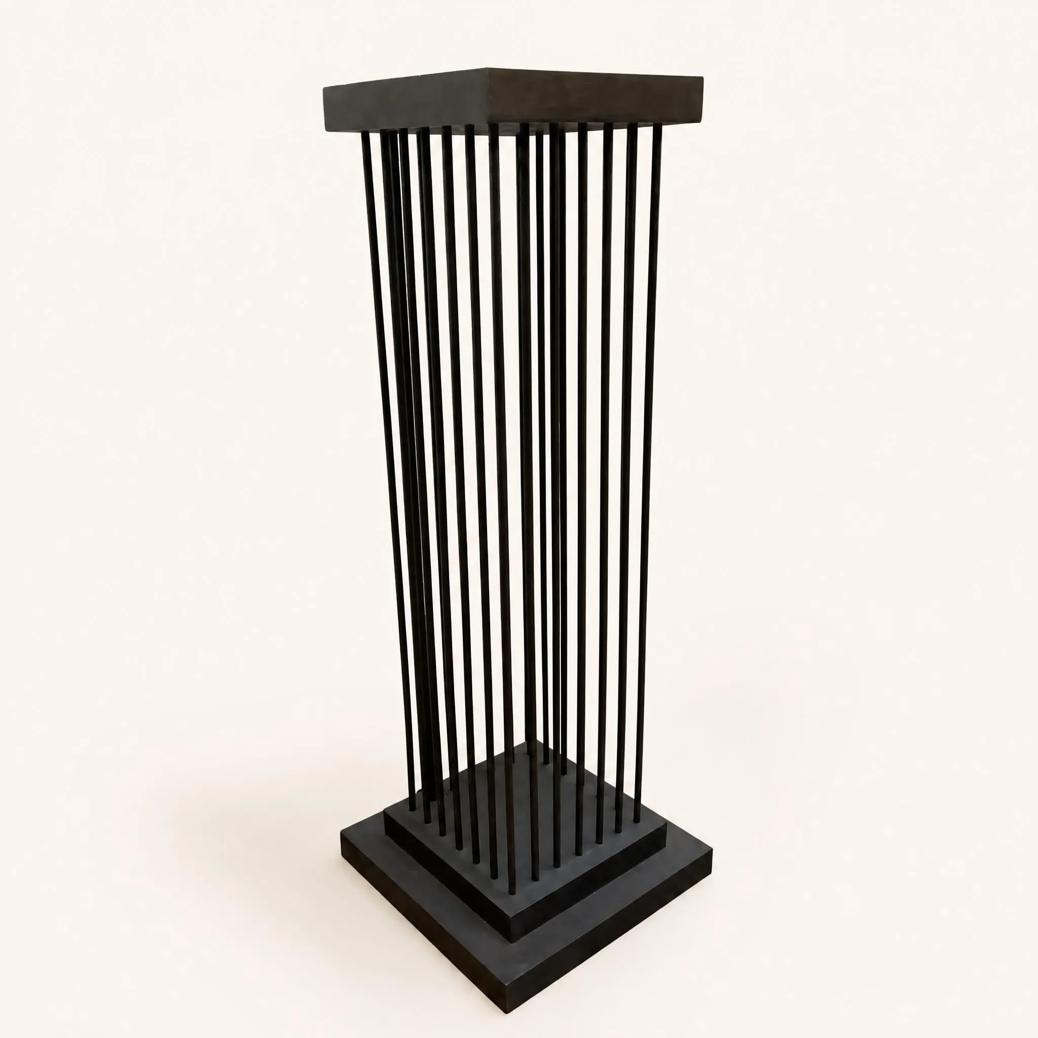 Gaine de présentation de section carrée en fer forgé patiné. Sa structure minimaliste en "cage" est constituée d'une grille périphérique de fines tiges verticales rectilignes. Celles-ci s'insèrent entre une base pleine à double ressaut (piétement à gradins) et un plateau supérieur carré destiné à recevoir un objet d'art. Le design rigoureusement géométrique joue sur la transparence et le rythme des lignes verticales.